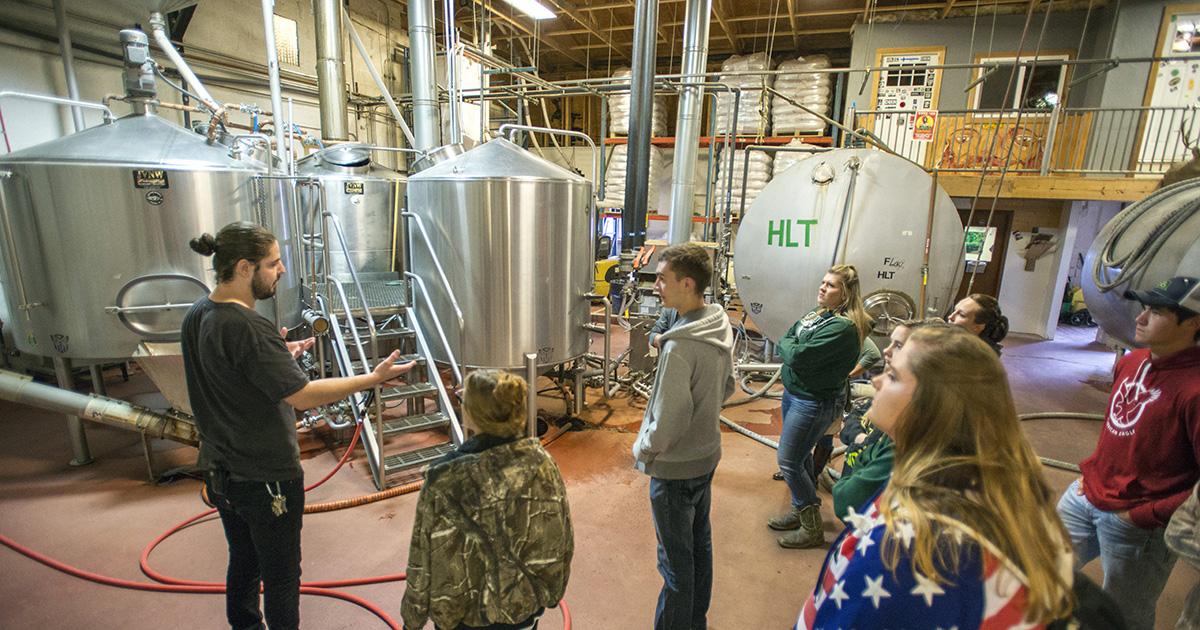 Students take a tour of Dark Horse Brewing Company with large metal tanks in the background.