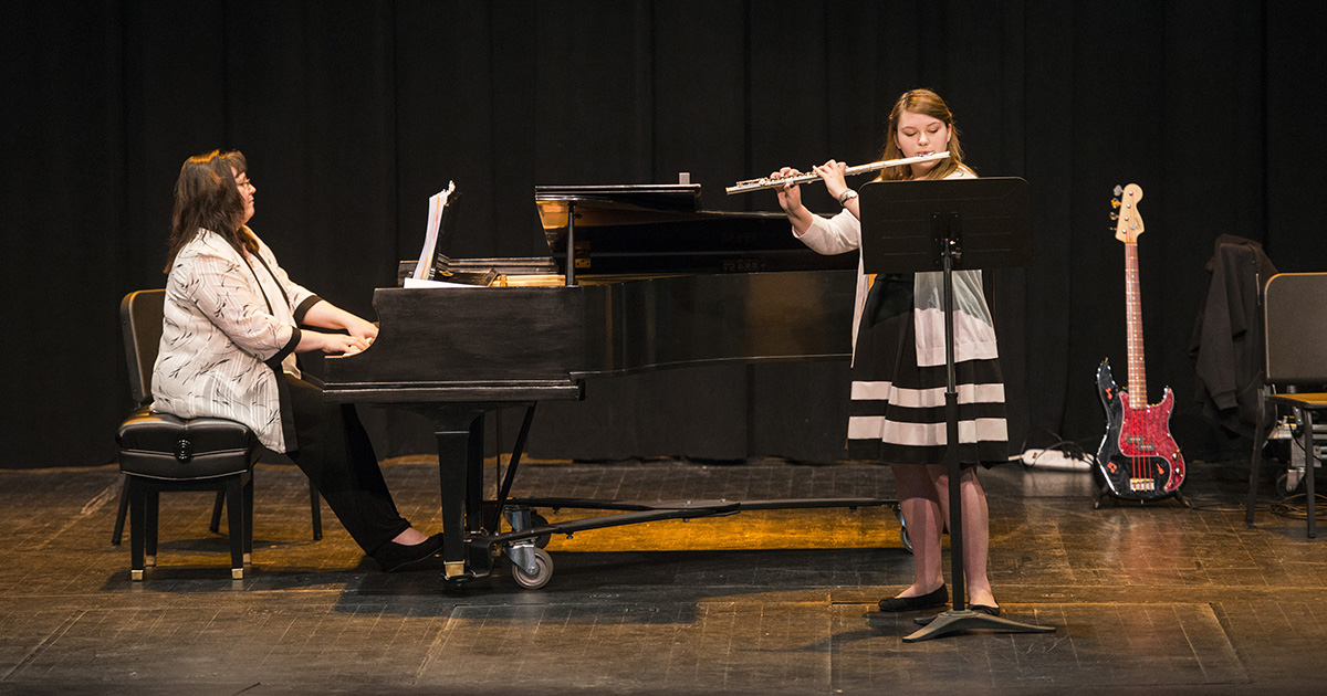 A student plays flute onstage while accompanied by a pianist.
