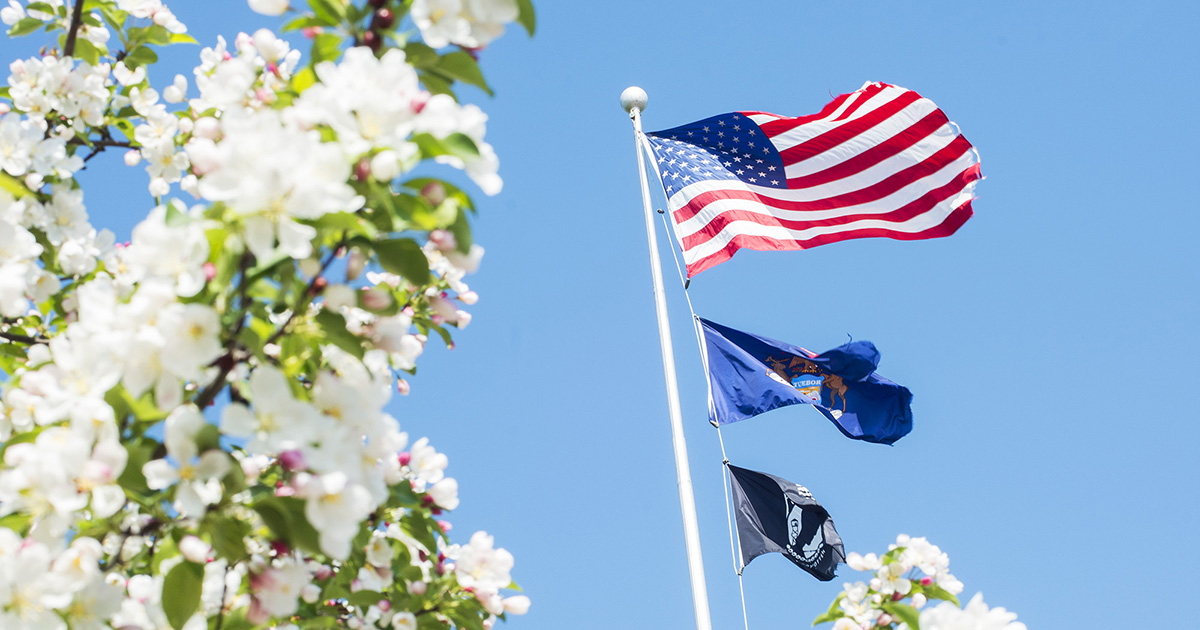 An American flag, Michigan state flag and POW flag fly with flowers in the foreground.