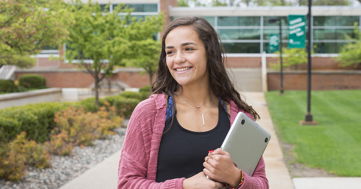 A student smiles while holding a laptop and walking outside on campus.