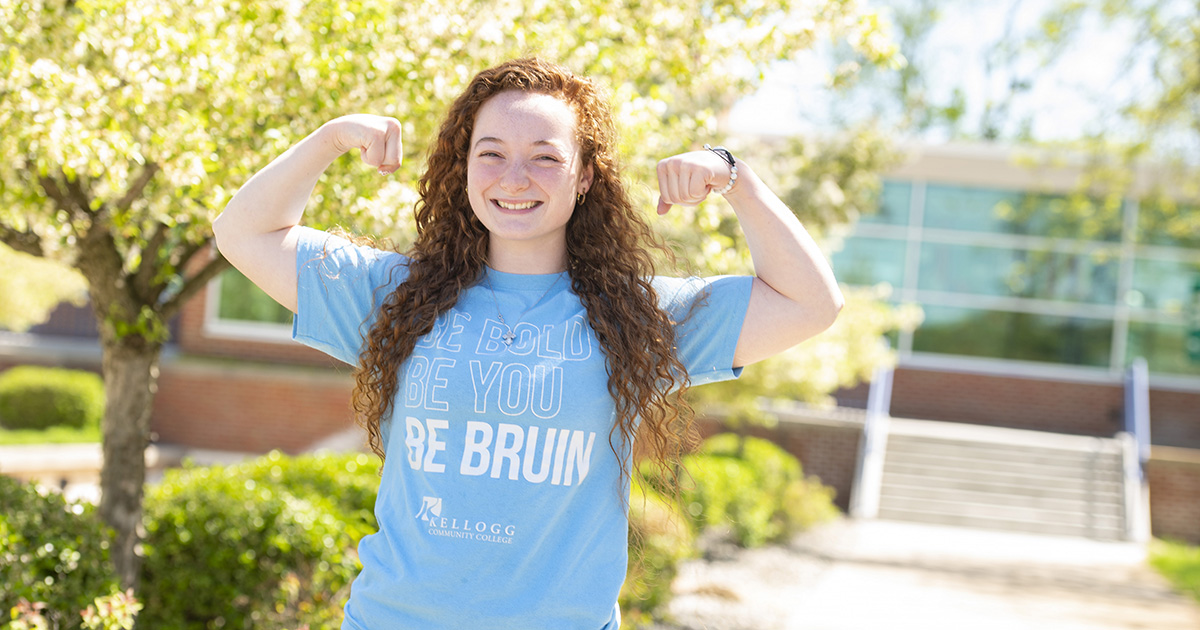 A female student smiles while flexing outside and wearing a light blue Be Bruin T-shirt.