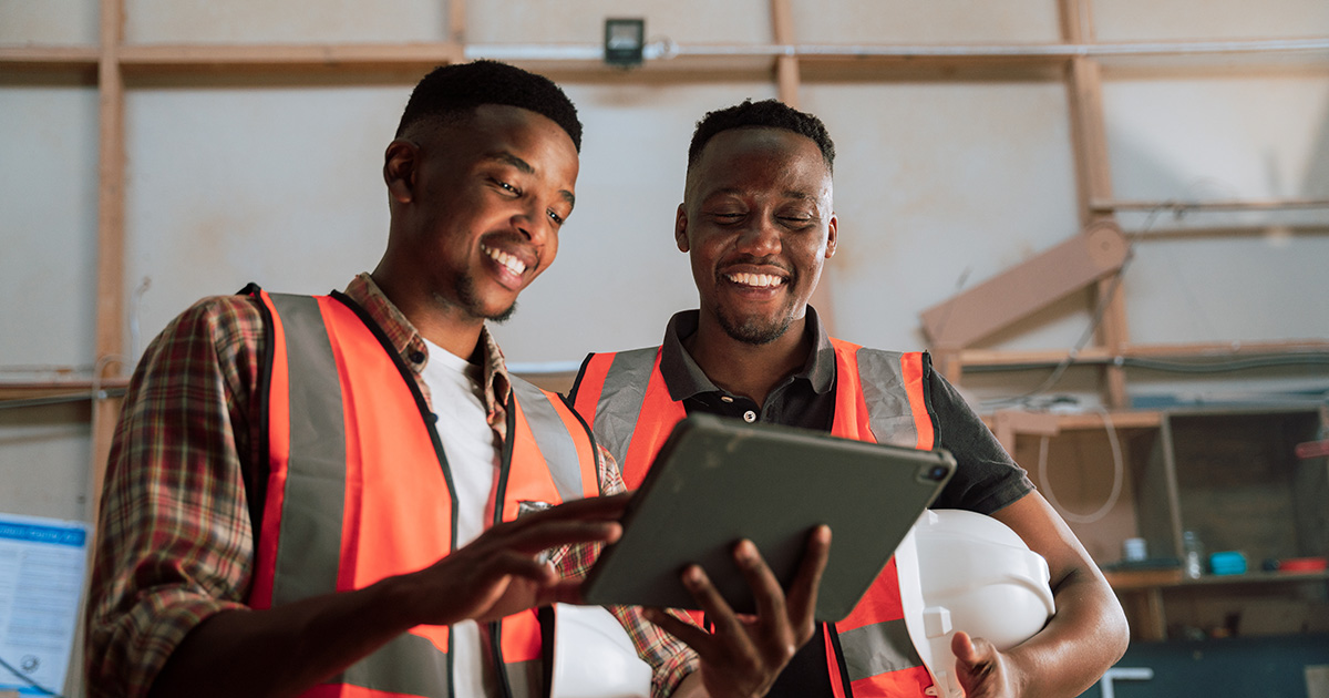 Two men in construction/safety outfits smile while looking at a tablet.